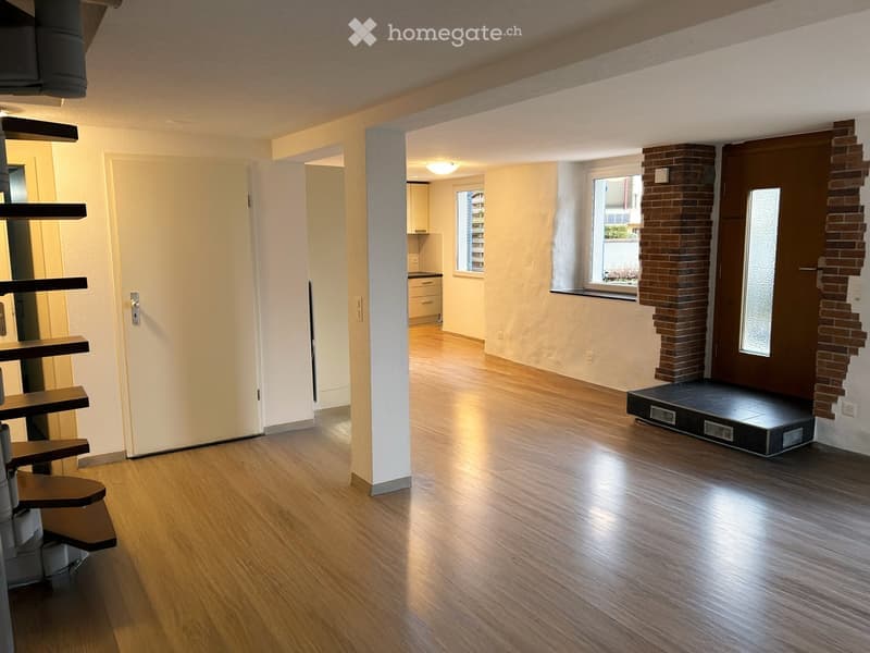 Empty room with wooden flooring, white walls, and a staircase leading to another level. The room has a large window with a view of the outside and a partially visible kitchen area in the background.
