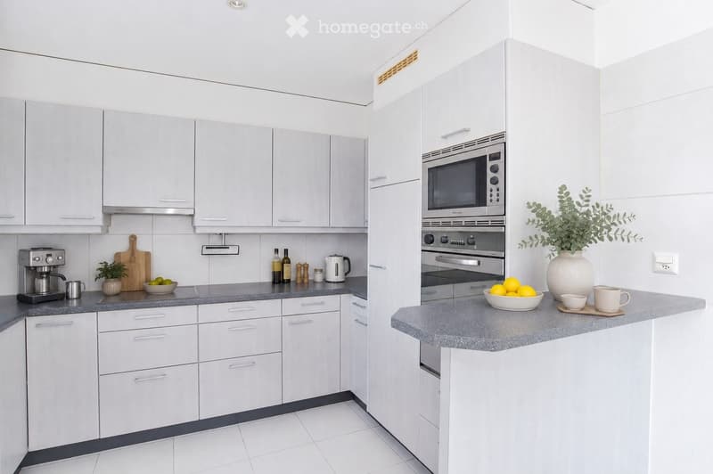 Modern kitchen with white cabinets, grey countertops, and stainless steel appliances. Includes a coffee maker, cutting board, bowl, oven, microwave, and decorative items on the counter.