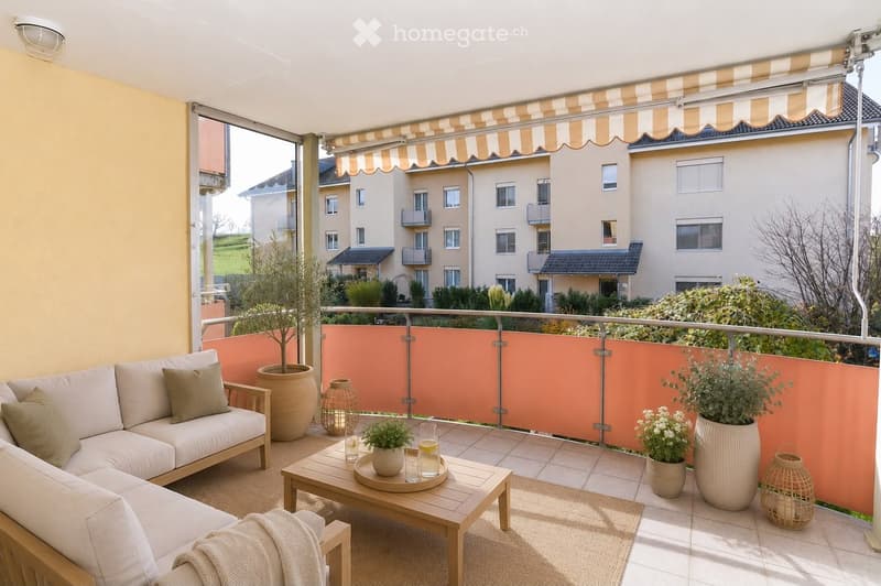 Balcony with a white sofa, wooden coffee table, potted plants, and a striped awning. The balcony features a modern design with a metal railing and a view of a residential area.