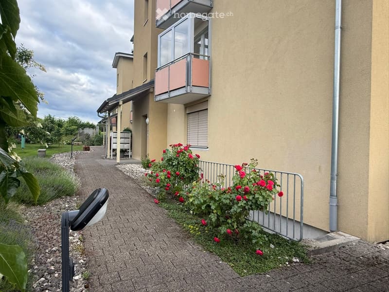 Modern multi-story residential building with beige facade, balcony, and well-maintained garden area with red flowers and shrubs.