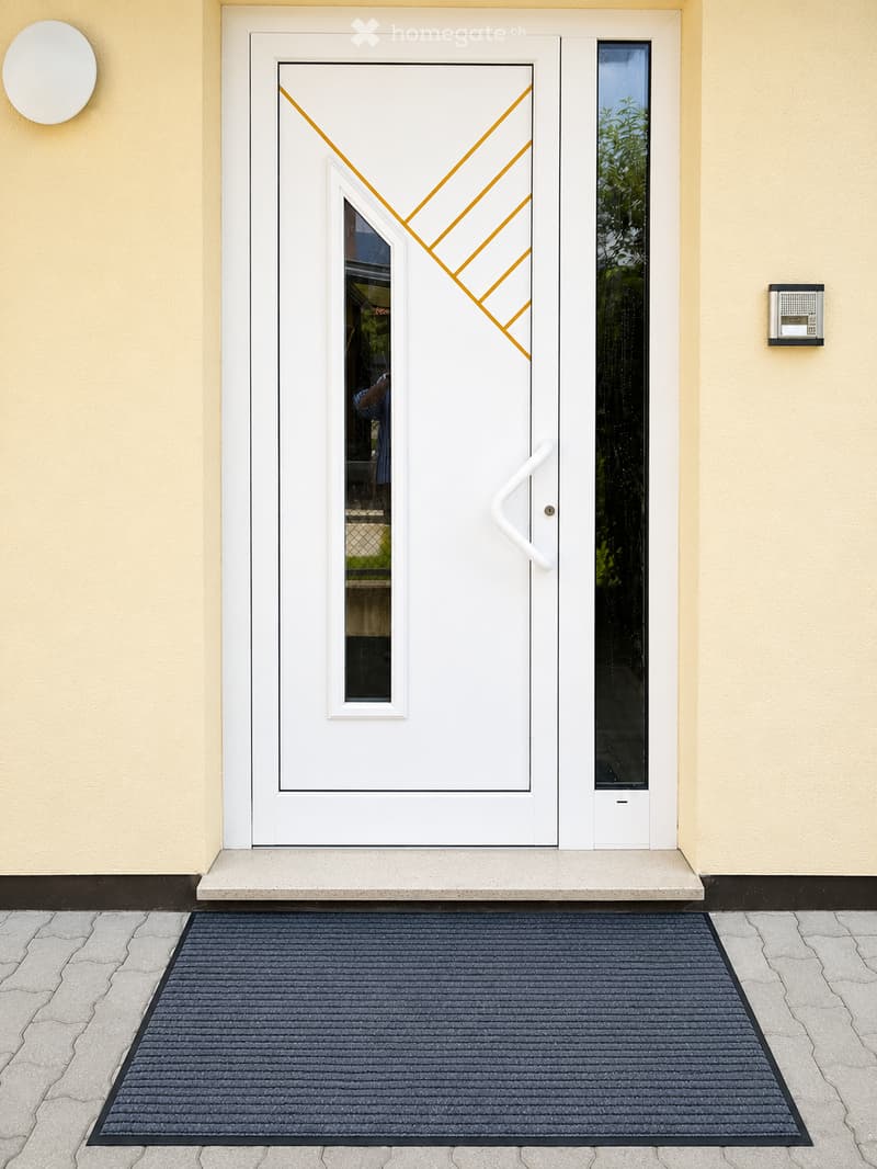 Front entrance door with a modern white frame and glass panels, featuring a decorative orange diagonal design on the glass. A black doormat is placed on the gray tiled ground in front of the door.