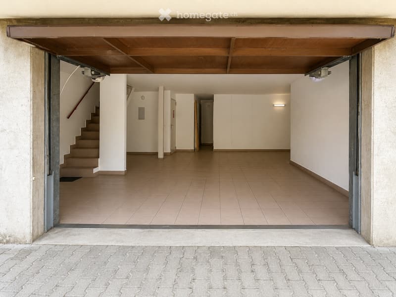 Empty hallway with tiled floor, white walls, and a staircase leading upwards. The area is well-lit with recessed lighting and has a wooden ceiling visible through an open doorway.