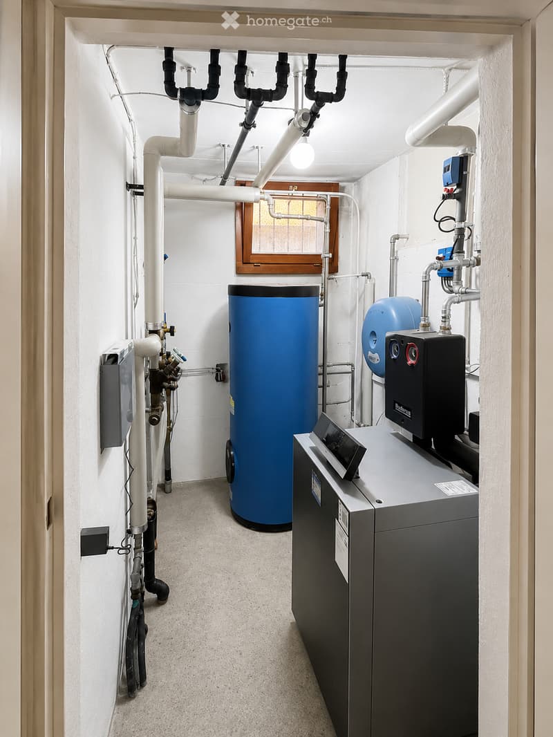 Utility room with white walls, grey floor, multiple pipes, water tank, control panel, and a small window with wooden frame.