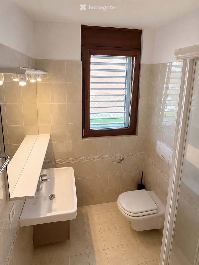 Modern bathroom with beige tiles, a white sink with a mirror, a toilet, and a window with blinds.