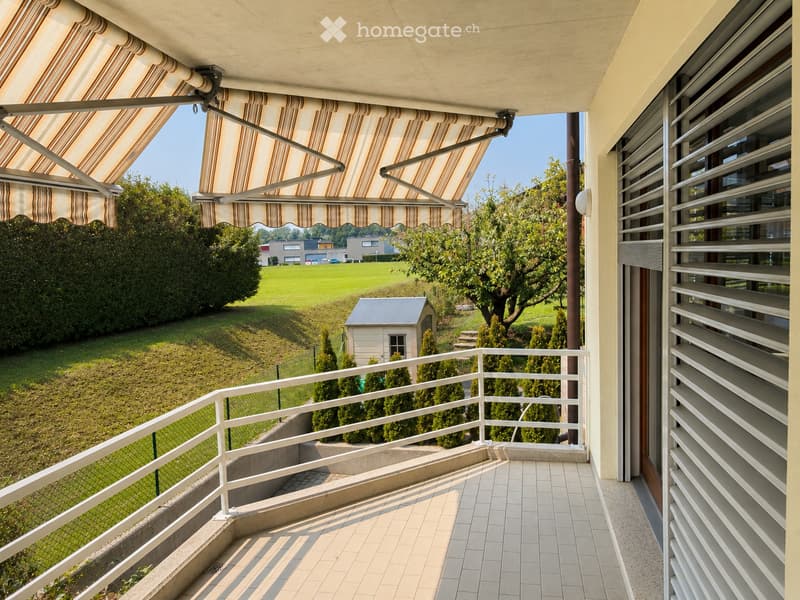 Balcony with tiled floor, white metal railing, striped awning, overlooking a green lawn and trees.