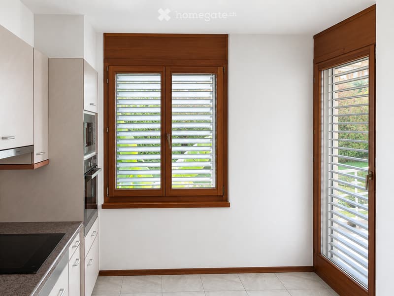 Modern kitchen with light-colored cabinets, stainless steel appliances, and large windows with wooden frames and blinds.