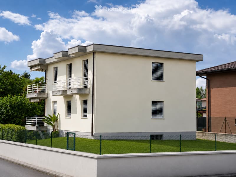 Two-story house with light beige exterior, multiple windows, balconies, and a fenced front yard.