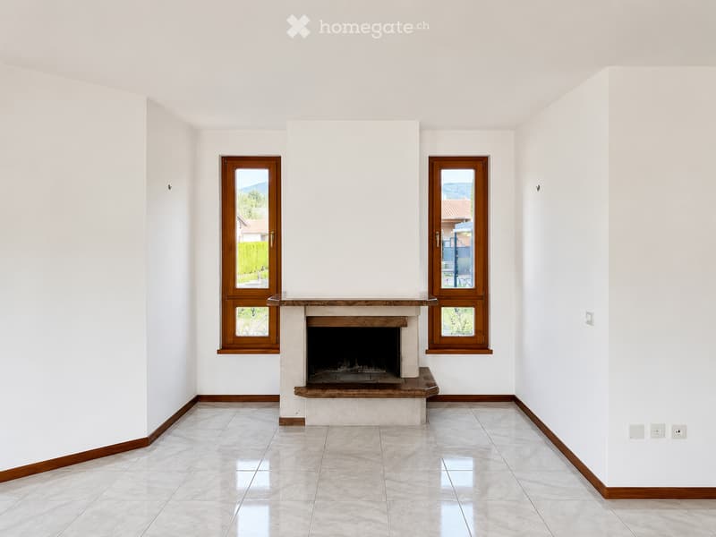 Empty room with white walls, tiled floor, fireplace, and two windows offering a view of greenery and buildings.