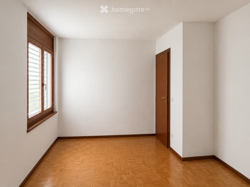 Empty room with light-colored walls, wooden herringbone flooring, and a single window with wooden frame and shutters. A closed wooden door is visible on the right side.