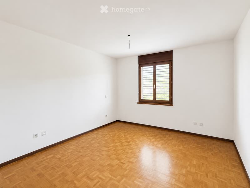 Empty room with white walls, wooden herringbone flooring, and a single window with blinds.