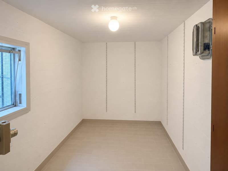 Empty room with white walls, beige tiled floor, and a single window with blinds. A light fixture is mounted on the ceiling, and a hand sanitizer dispenser is mounted on the wall.
