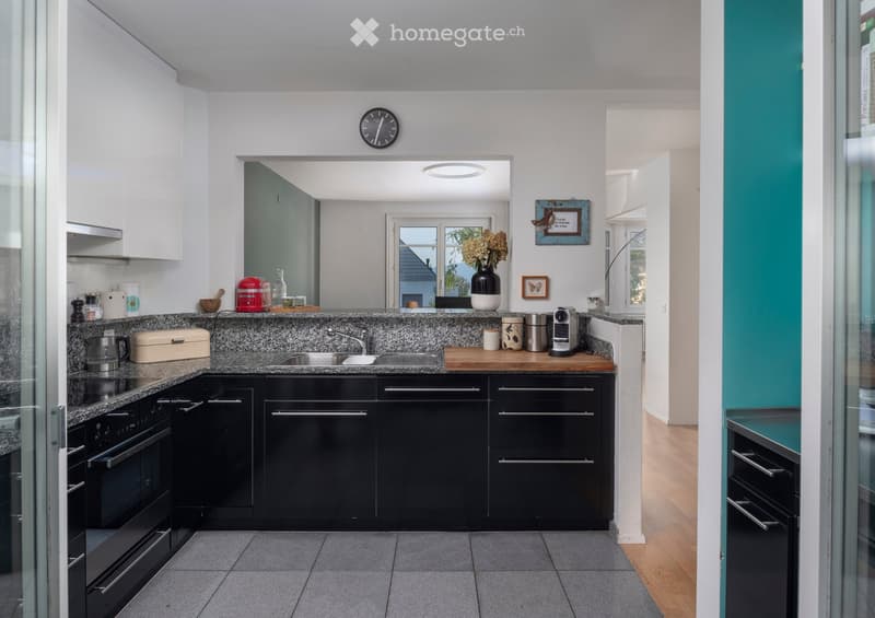 Modern kitchen with black cabinets, granite countertops, stainless steel appliances, and a central island with a sink and faucet. The space features a clock on the wall, a vase with flowers, and various kitchen items on the counter.
