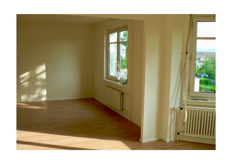 Empty room with wooden flooring, white walls, and large windows allowing natural light. Radiators are visible under the windows.