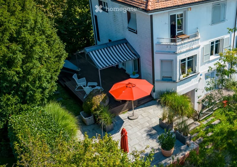 A modern two-story house with a large outdoor patio area, featuring potted plants, an orange umbrella, and a covered seating area with white chairs.