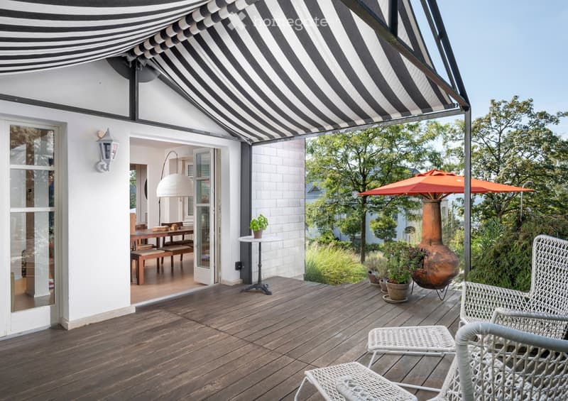 Wooden deck with white wicker chairs, small table, outdoor umbrella, and a large ceramic vase. The deck is covered by a black and white striped awning, with a view of a garden and trees.