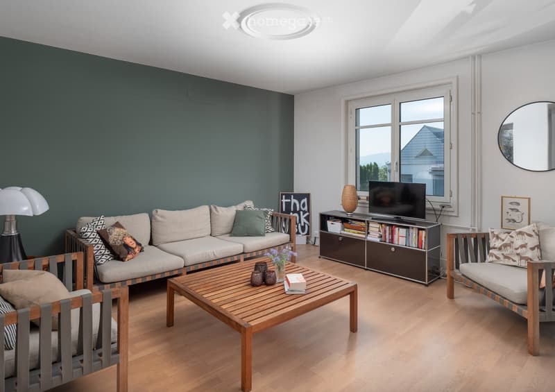 Modern living room with light wooden flooring, grey sectional sofa, wooden coffee table, and a TV stand with books and decorative items.