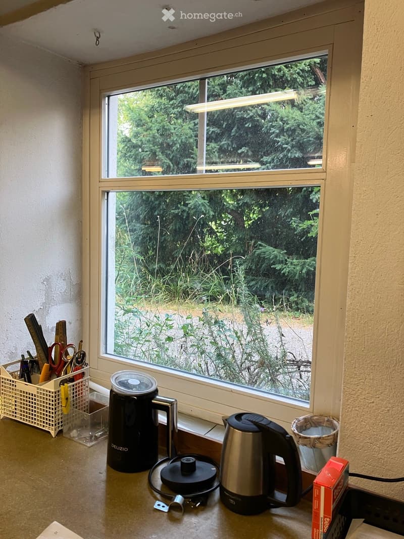 A small kitchen area with a window overlooking a garden, featuring a coffee maker, kettle, basket, and various small items on the counter.