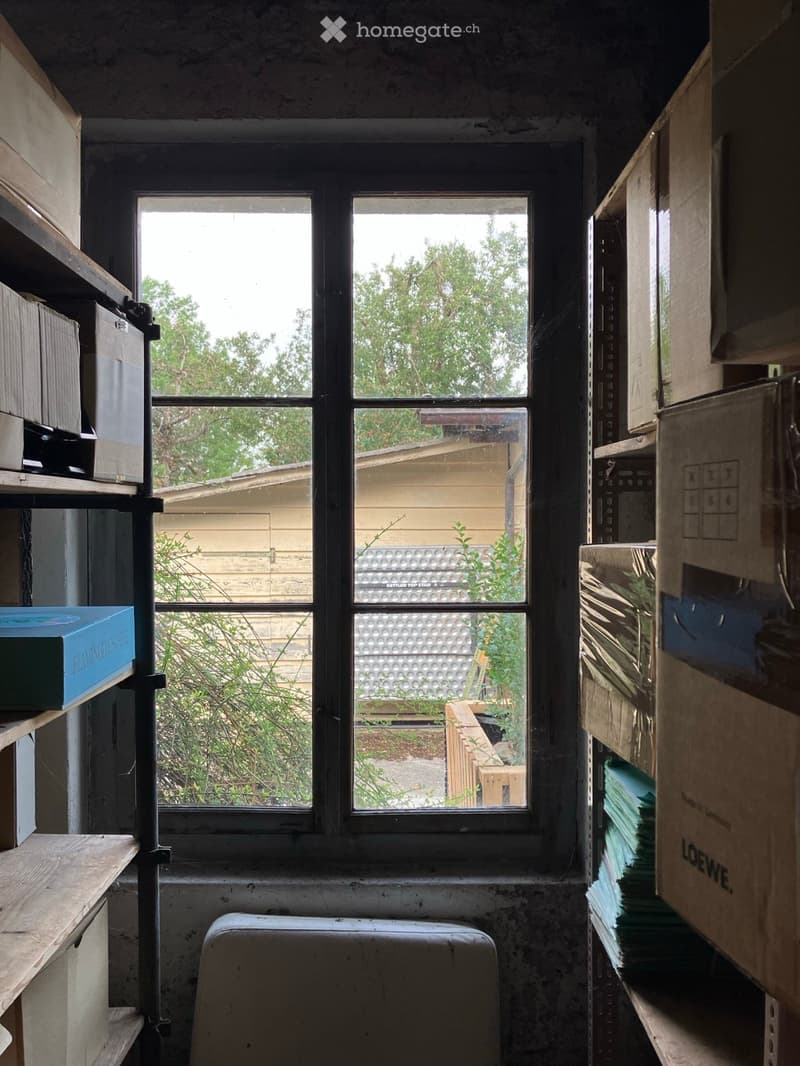 Empty room with exposed concrete walls, a large window showing outdoor view, metal shelving units with boxes, and a small wooden platform.