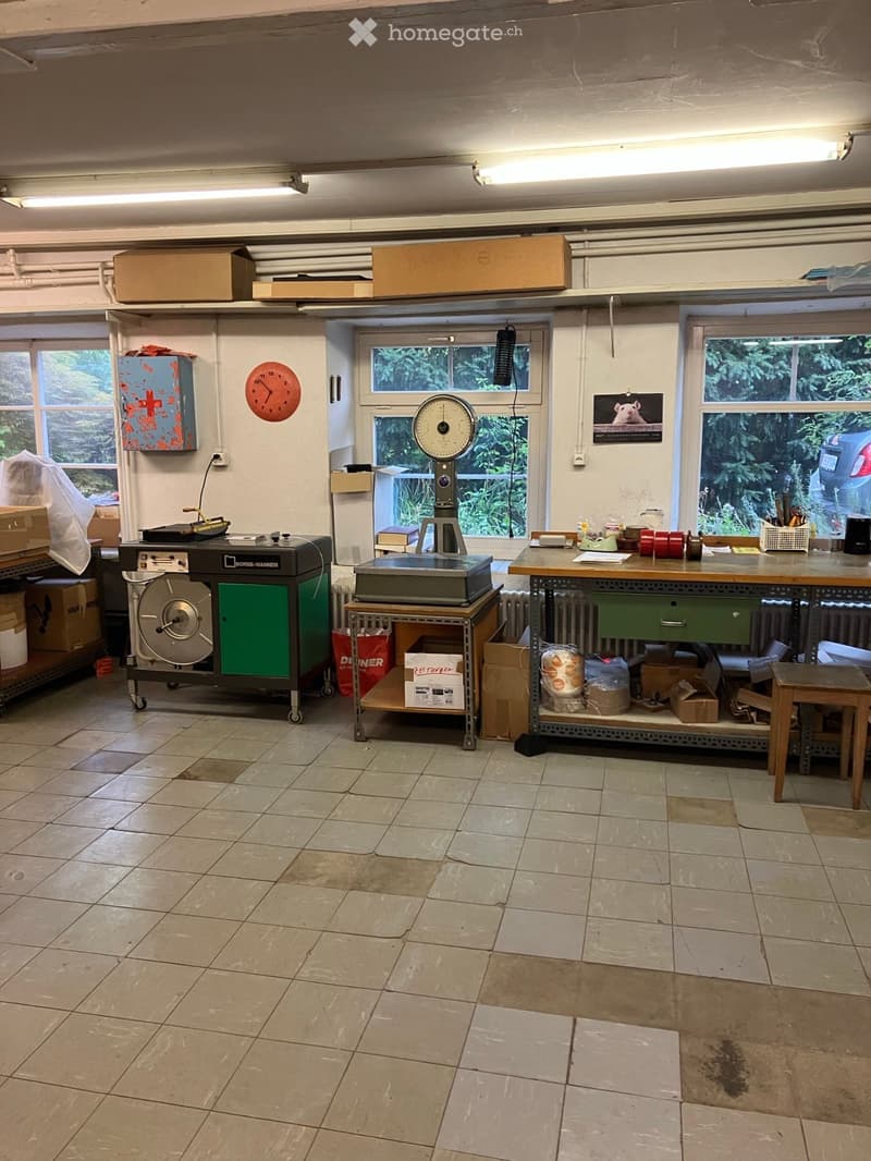 Workshop or storage area with tiled floor, large windows, table with various items, scale, cabinet, clock, and radiator.