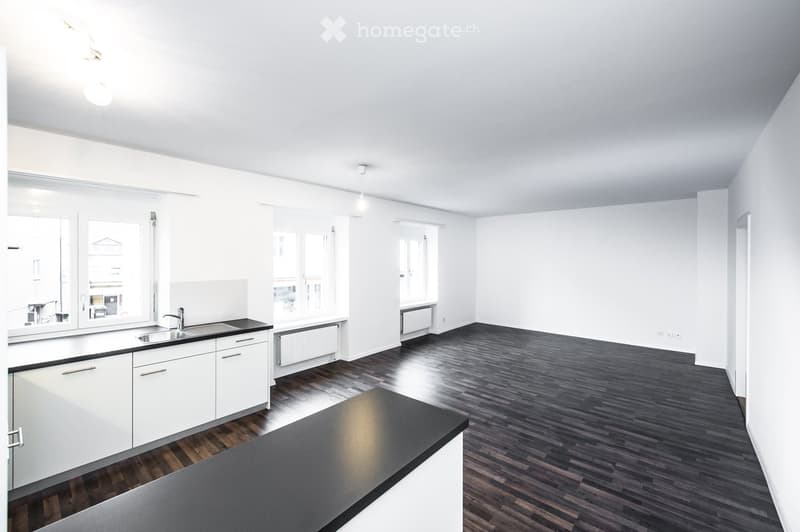 Empty room with white walls, dark wooden flooring, and a small kitchen area featuring white cabinets and a sink.