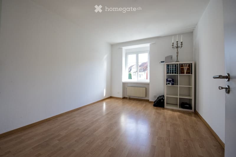 A bright, unfurnished room with wooden flooring, white walls, and a large window letting in natural light. A small shelf with books and a candelabra is visible, along with a radiator under the window and a couple of bags on the floor.