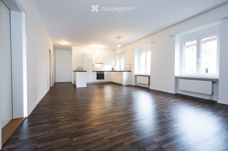 Empty room with wooden flooring, white walls, and large windows. The room features a kitchen area with white cabinets and a countertop, but no furniture or personal items are present.