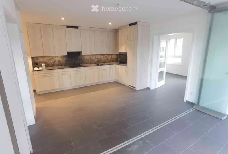 Modern kitchen with light wooden cabinets, stainless steel appliances, and a dark tiled floor. The space is open and bright with large windows.