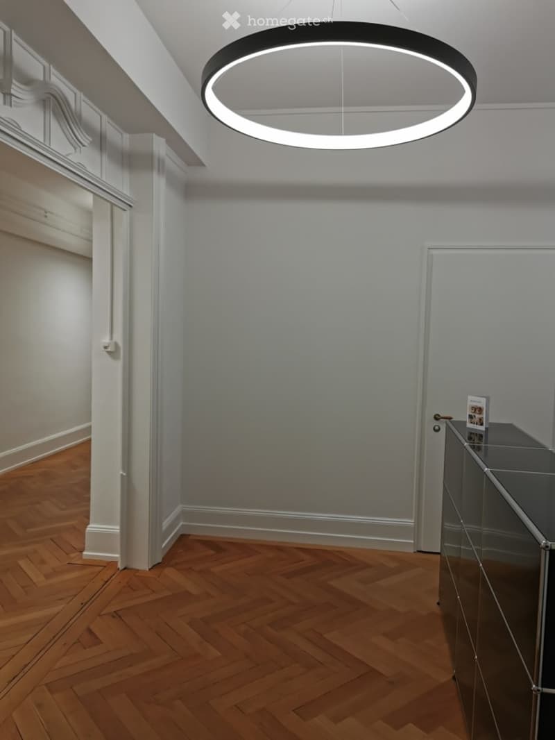 Empty room with wooden floor, white walls, large circular light fixture, and a cabinet with a picture frame on top