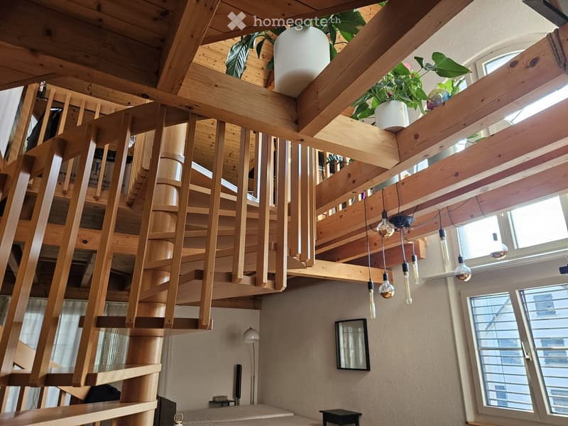 Spacious, modern living area with a wooden spiral staircase leading to an upper level, exposed wooden beams, pendant lights, potted plants, a mirror, and a small table and stool near the window.