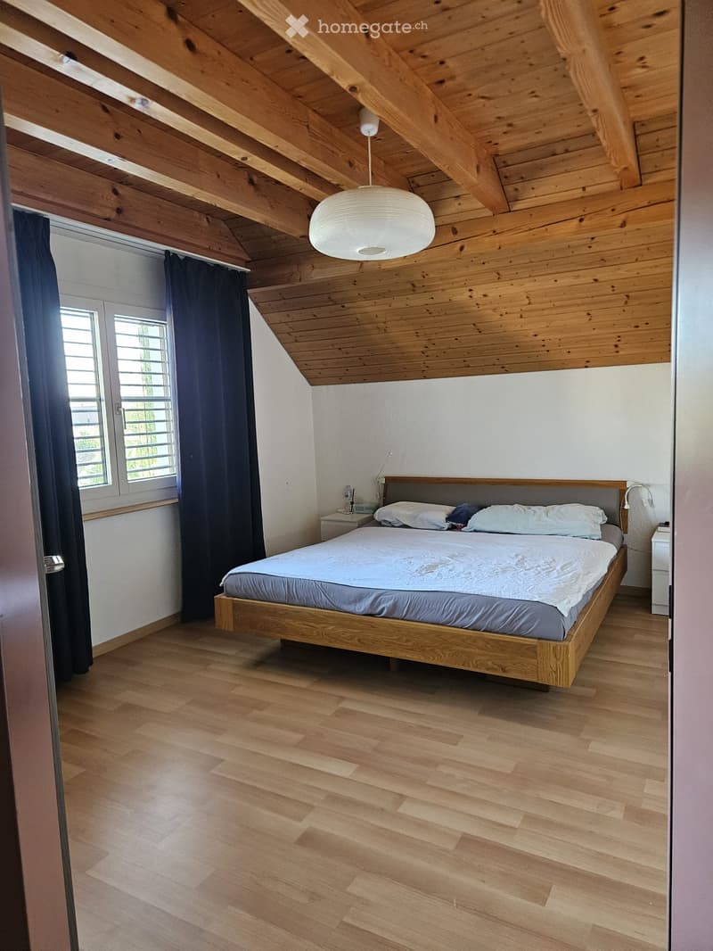 Cozy bedroom with wooden floors, slanted wooden ceiling, large window with dark curtains, and a modern wooden bed with white bedding.