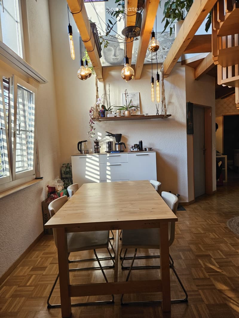 Bright, modern kitchen and dining area with wooden flooring, pendant lights, white cabinets, a wooden dining table with chairs, and a coffee maker on the counter.