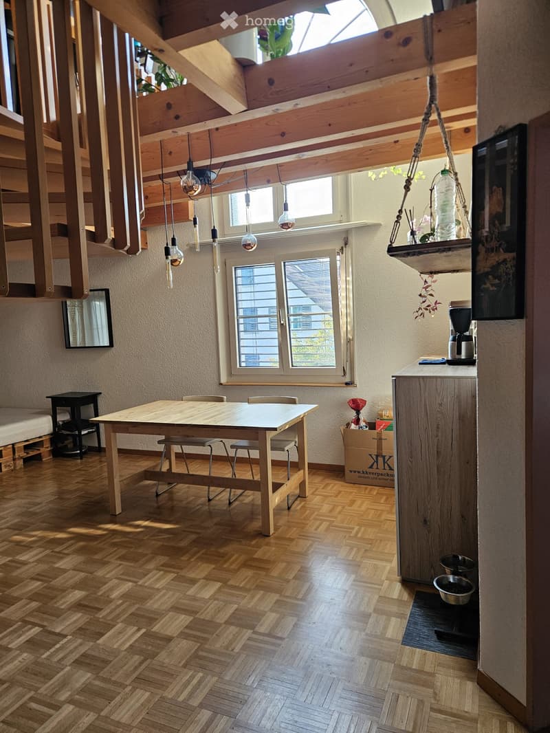 Bright, open room with wooden herringbone flooring, exposed wooden beams, and large windows. Features a dining table with chairs, a kitchen counter with a coffee maker, and a hanging plant. A small black table and mirror are visible on the left side.