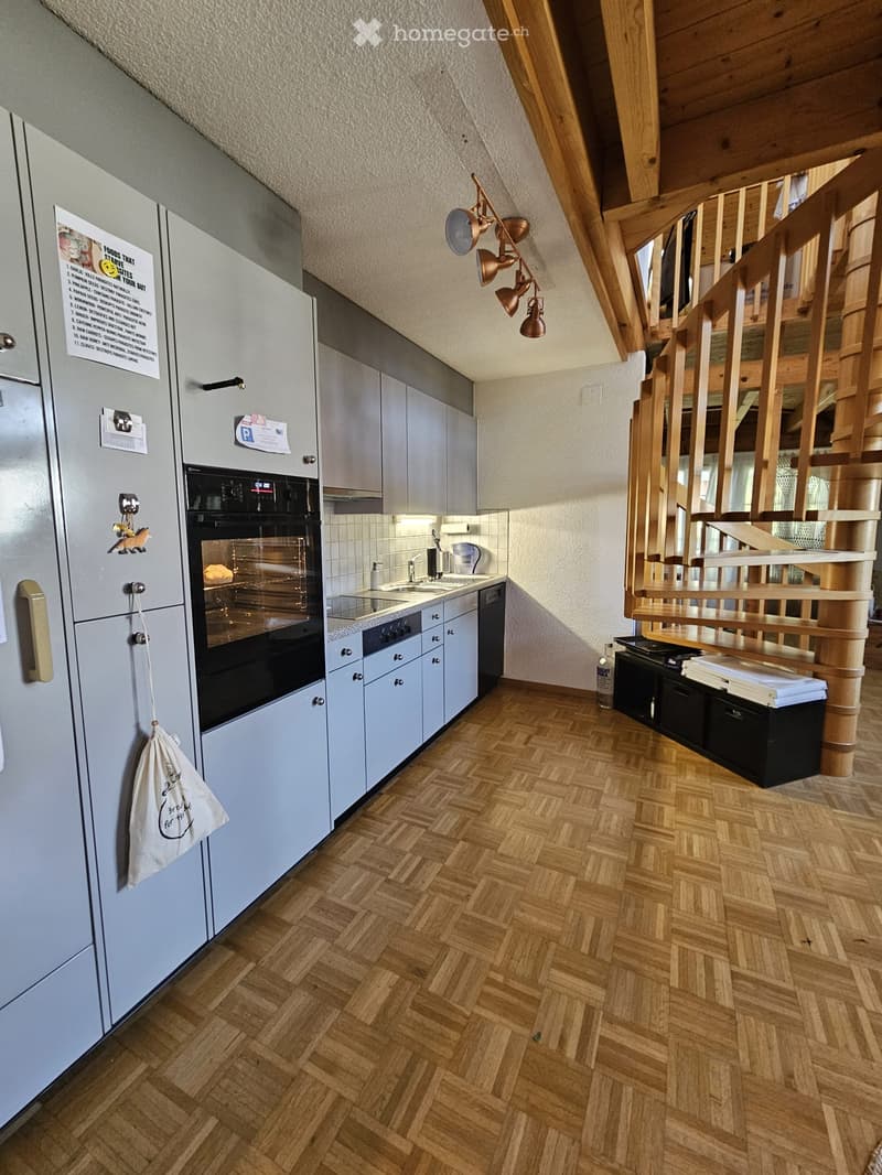 Modern kitchen with light grey cabinets, stainless steel appliances, and a wooden herringbone floor. Features a refrigerator, oven, sink, and a hanging light fixture.
