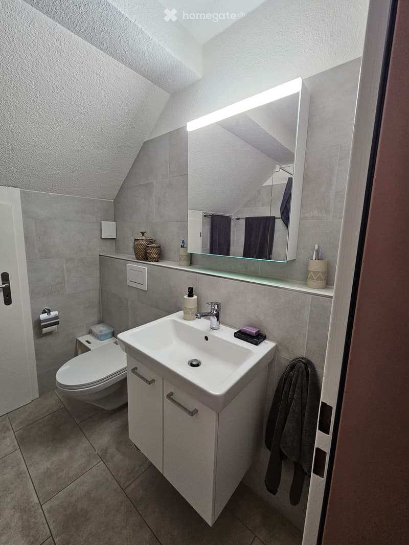 Modern bathroom with a white sink, chrome faucet, toilet, large mirror with built-in lighting, and decorative baskets on the counter.
