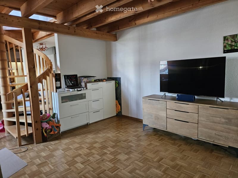 Living room with wooden floor, exposed wooden beams, television on a wooden cabinet, laptop on a white cabinet, staircase with wooden railing, and a wine bottle on the shelf.
