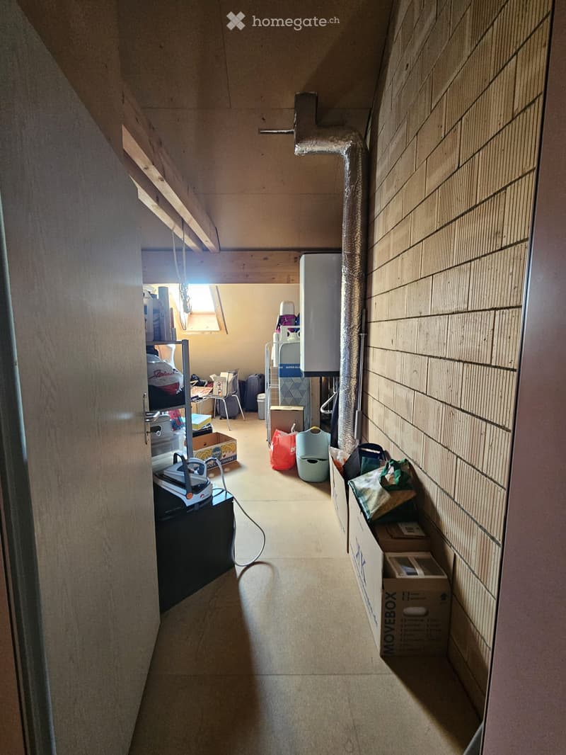 A narrow hallway with exposed wooden beams and tiled walls, featuring storage boxes, a water tank, and various household items along the walls.