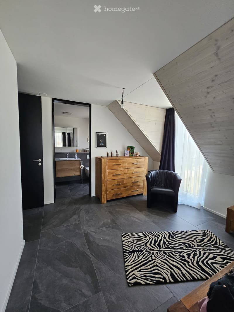 A modern, furnished room with a slanted wooden ceiling, dark grey tiled floor, a black leather armchair, a wooden chest of drawers, a zebra print rug, a framed picture, and an adjacent bathroom with a sink and toilet.