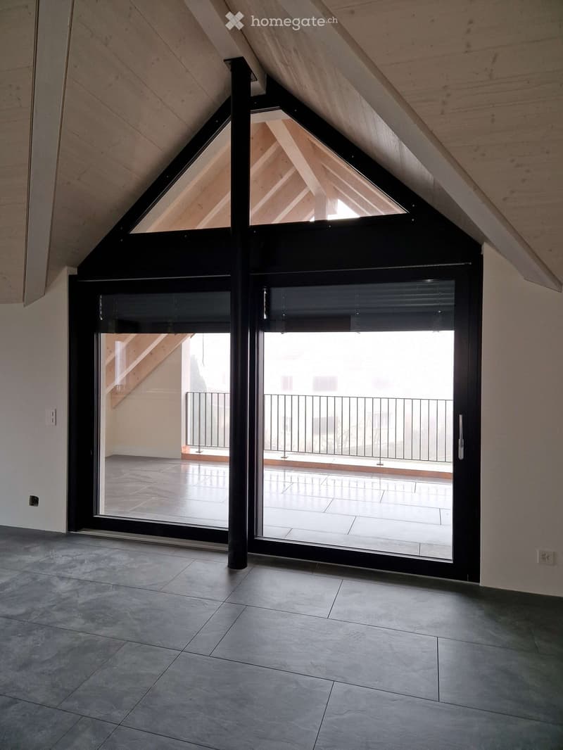 An empty attic space with exposed wooden beams, large sliding glass doors leading to a balcony, and a tiled floor.