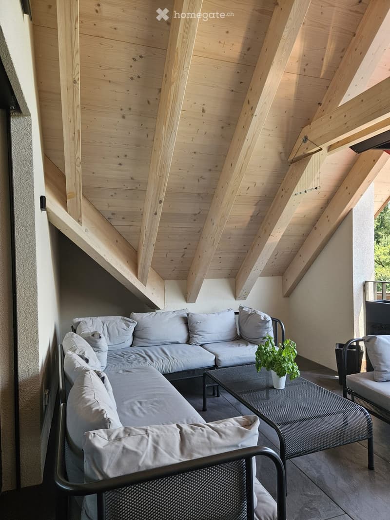 A modern, wooden-slatted balcony with a grey sectional sofa, a small table with a potted plant, and a matching chair.