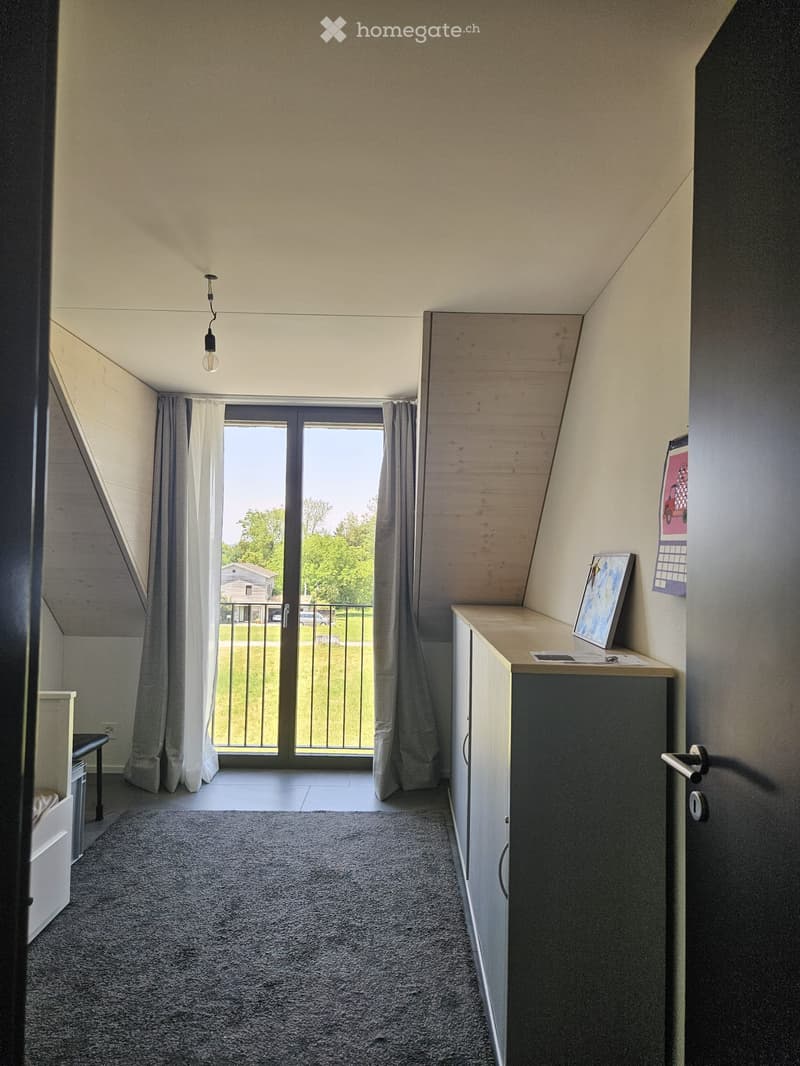 A small, modern room with a slanted ceiling, carpeted floor, and large window with curtains leading to a balcony. Includes a small cabinet, table, chair, and picture frame.