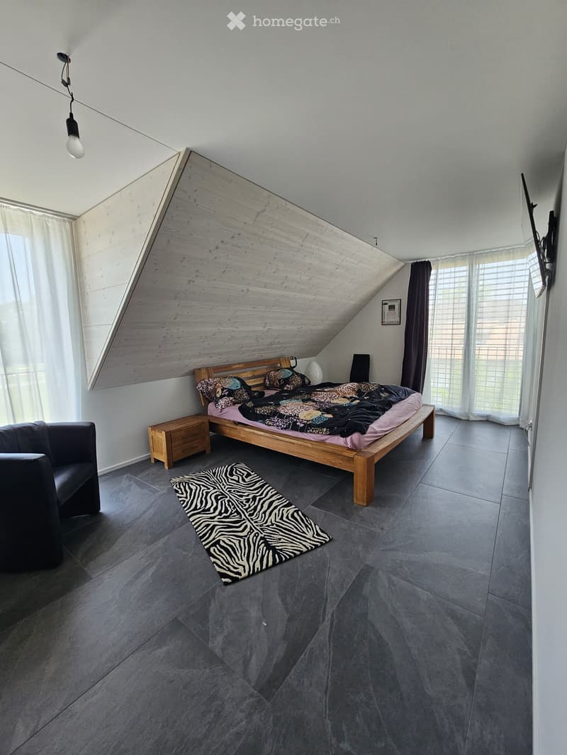 Modern bedroom with a wooden bed, nightstand, black chair, and zebra-print rug on a grey tiled floor. Large windows with white curtains and dark curtains on one side.