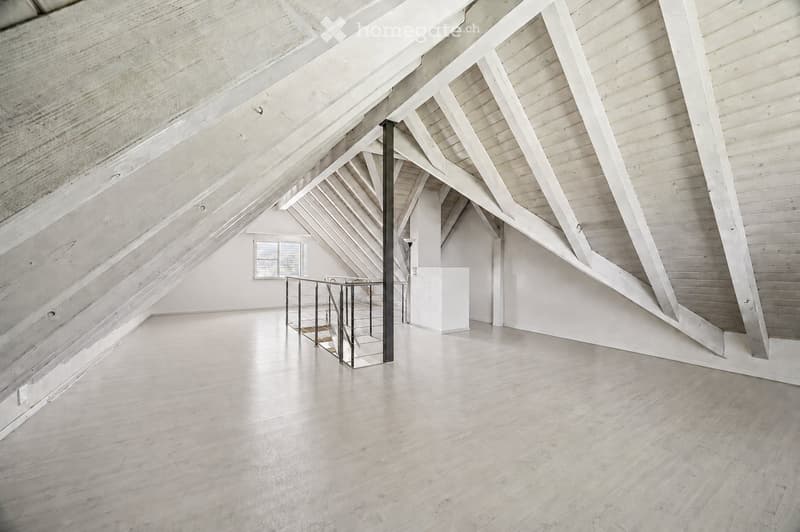 Open, airy attic space with exposed wooden beams and a modern metal staircase leading to an upper level. The room features a large skylight window and a polished wooden floor.