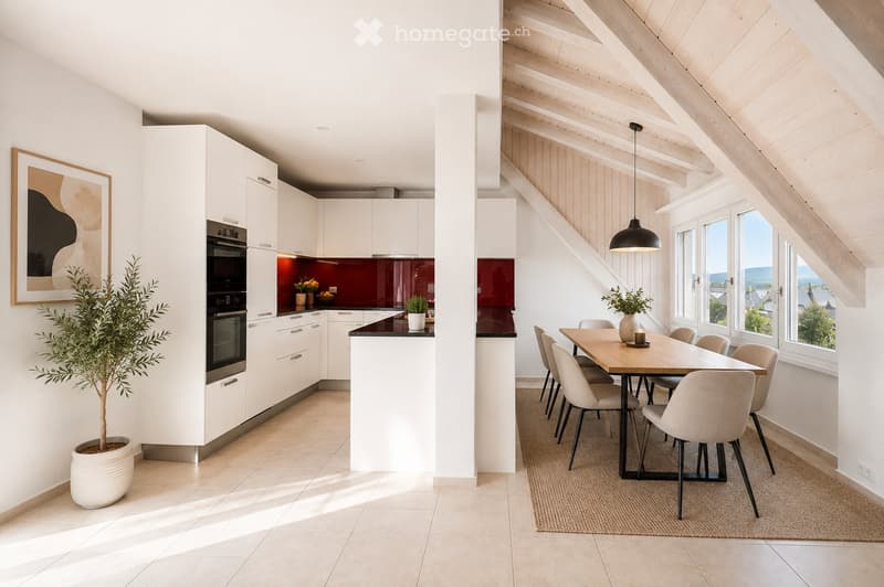 Modern kitchen with white cabinets, a built-in oven, a central island, and a dining area with a wooden table and chairs. The space features large windows with a scenic view, a hanging pendant light, and a potted plant near the kitchen island.