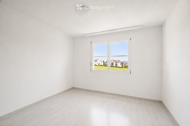 Empty room with white walls, light wooden flooring, and large windows offering a view of a green area and buildings.