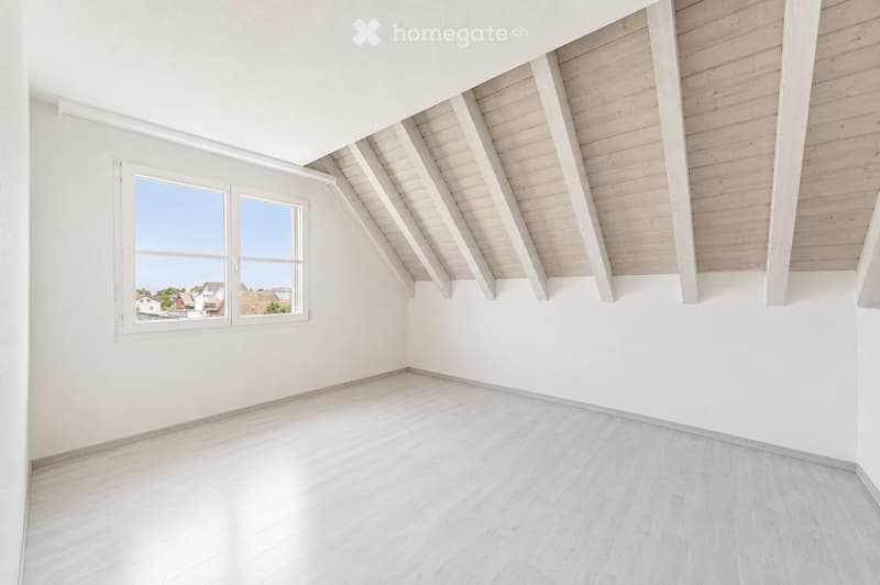 Empty room with white walls, light wooden flooring, and a slanted wooden ceiling. A large window offers a view of the outside.