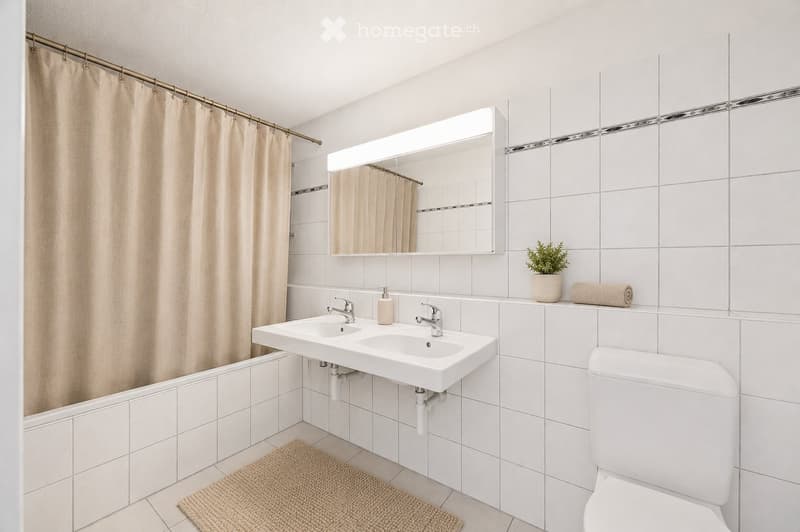 Modern bathroom with white tiles, double sink, toilet, large mirror with built-in lighting, shower curtain, small plant, and towel on the counter.