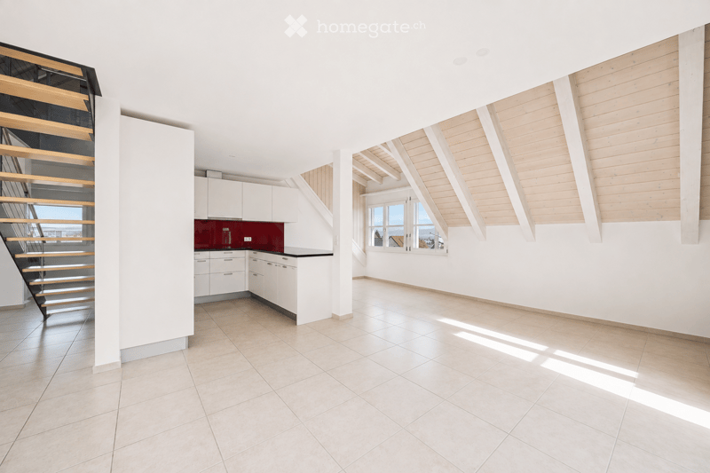 Bright, open-plan space with tiled flooring, white walls, and wooden beams on the ceiling. Features a modern staircase with glass railings and a small kitchen area with white cabinets and a red backsplash.