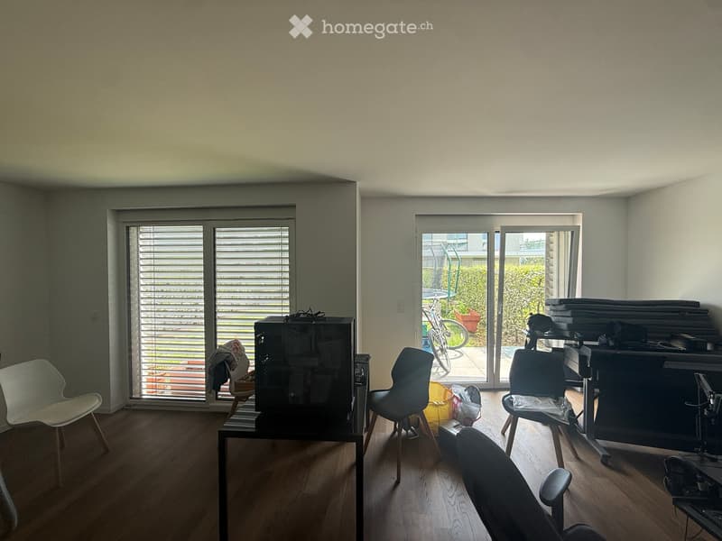 A modern, well-lit room with wooden flooring, large windows, and sliding glass doors leading to a patio. The room features several chairs, a table, a screen, and a laptop, with a bicycle visible outside the sliding doors.