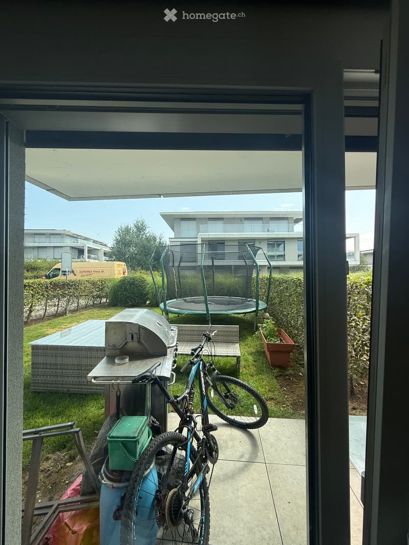 Balcony with a bicycle, a small table, a bench, a trash bin, a potted plant, and a trampoline in the background.