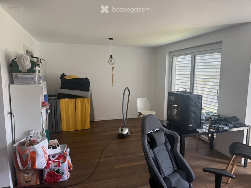A furnished room with a gaming chair, desk, and various items including a refrigerator, a lamp, and a vacuum cleaner. The room has wooden flooring and large windows with blinds.
